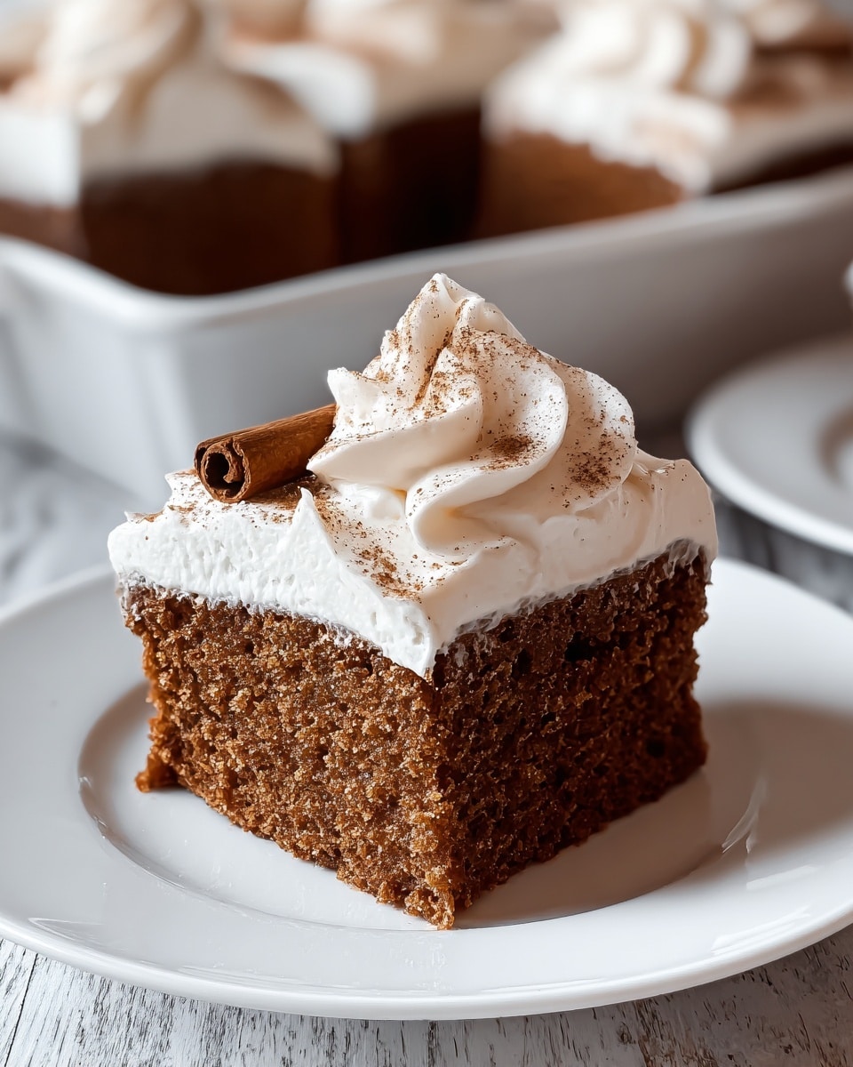 This image shows a single slice of brown cake with a smooth texture, placed on a white plate. The cake has one thick layer with a dense, moist inside. On top, there is a thick layer of creamy white frosting spread evenly, with a swirl of whipped cream in the center, sprinkled lightly with fine brown powder. A cinnamon stick lies diagonally on the frosting, adding decoration. In the background, more slices of the same cake appear blurred, resting together in a white dish on a white marbled surface. photo taken with an iphone --ar 4:5 --v 7