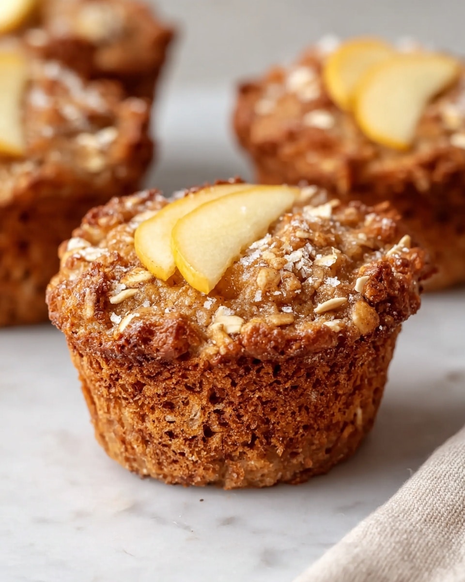 A close-up of a single muffin on a white marbled texture, showing a rough, golden-brown oat and cinnamon crust surrounding the muffin's light brown, textured interior. On top, there are two pale yellow apple slices partially embedded, sprinkled with coarse sugar crystals and fine cinnamon powder, and small oat flakes scattered around the surface. In the background, two more muffins are slightly blurred, keeping the attention on the front muffin. The corner of a beige cloth is visible at the bottom right photo taken with an iphone --ar 4:5 --v 7