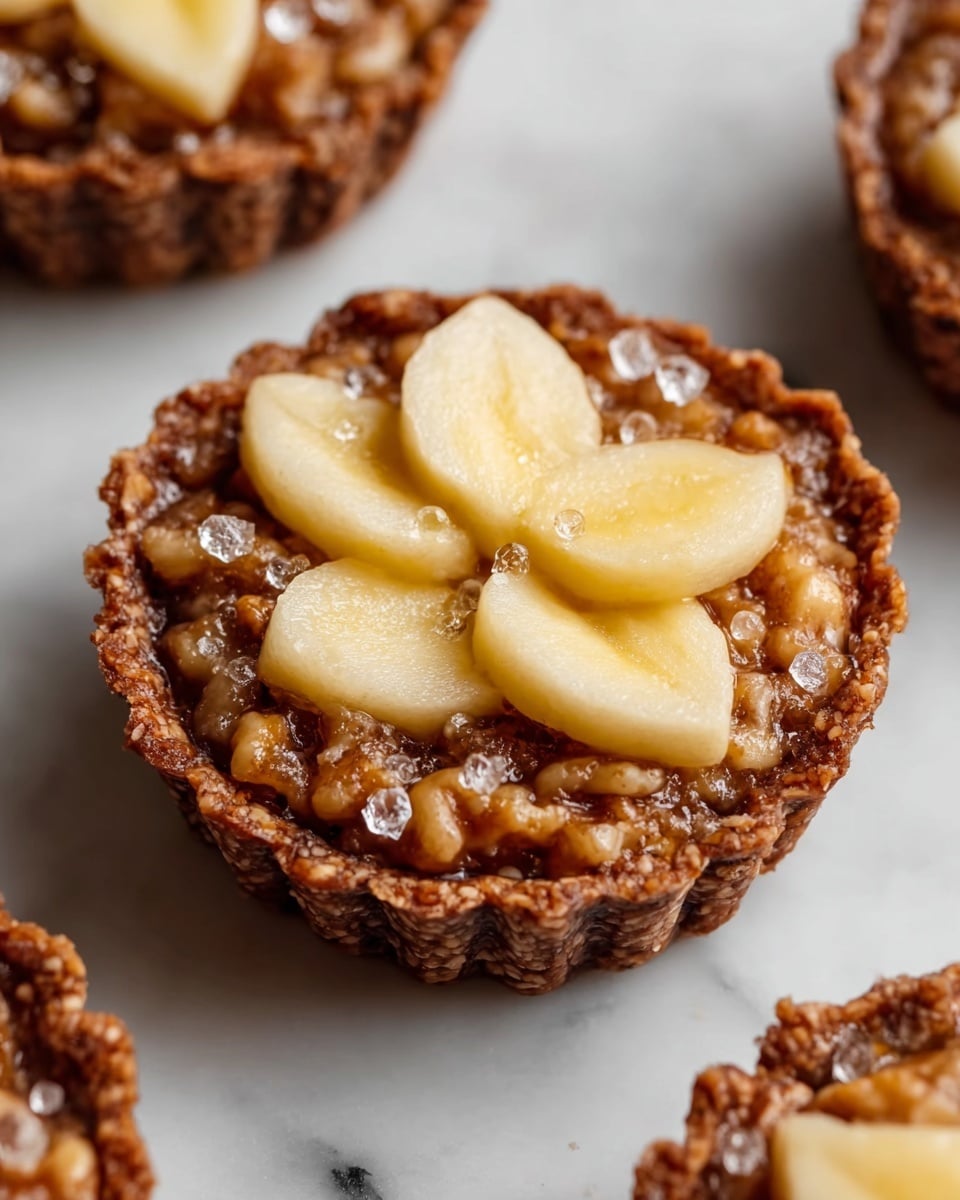 The image shows a close-up of a single mini tart on a white marbled surface, with parts of other tarts visible around it. The tart has a brown, crinkled crust with a rough texture. Inside the crust, there is a thick filling made of small walnut pieces held together by a sticky, glossy, brownish mixture that looks spiced with specks of cinnamon. On top, four pale yellow slices of fruit with a slightly rough surface are placed evenly, forming an opening in the center. The middle of the tart is decorated with small, clear sugar crystals adding a sparkling effect. Photo taken with an iphone --ar 4:5 --v 7
