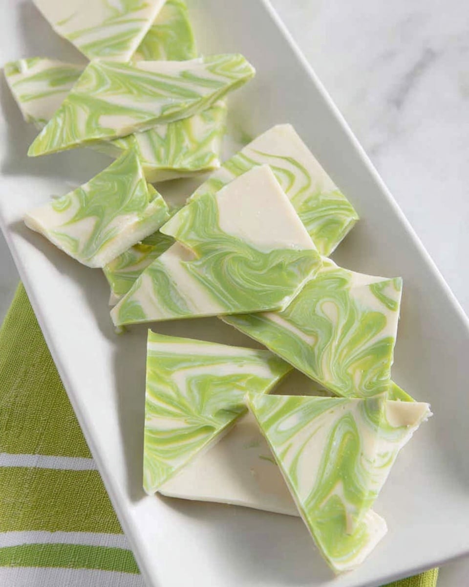 The image shows a white rectangular plate holding several pieces of swirl patterned candy bark. The bark is made of two layers; the top layer is a mix of light green and white swirls, creating a marbled effect with smooth curving lines, while the bottom layer is a solid white with a smooth texture. The pieces are broken into irregular shapes, some triangular, some more angular, neatly spread across the plate. This plate is set on a white marbled surface with a green and white striped cloth visible at the edges. photo taken with an iphone --ar 4:5 --v 7