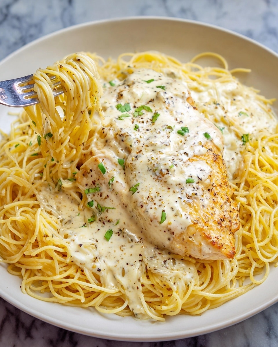 A round white plate holds a bed of thin yellow spaghetti noodles as the base layer, topped with a creamy white sauce mixed into the noodles. On top of the noodles, there is a large piece of golden-brown grilled chicken breast covered with creamy sauce, sprinkled with black pepper and small green herb pieces. A silver fork is picking up a small twisted bundle of noodles from the plate. The plate is set on a white marbled surface. Photo taken with an iphone --ar 4:5 --v 7
