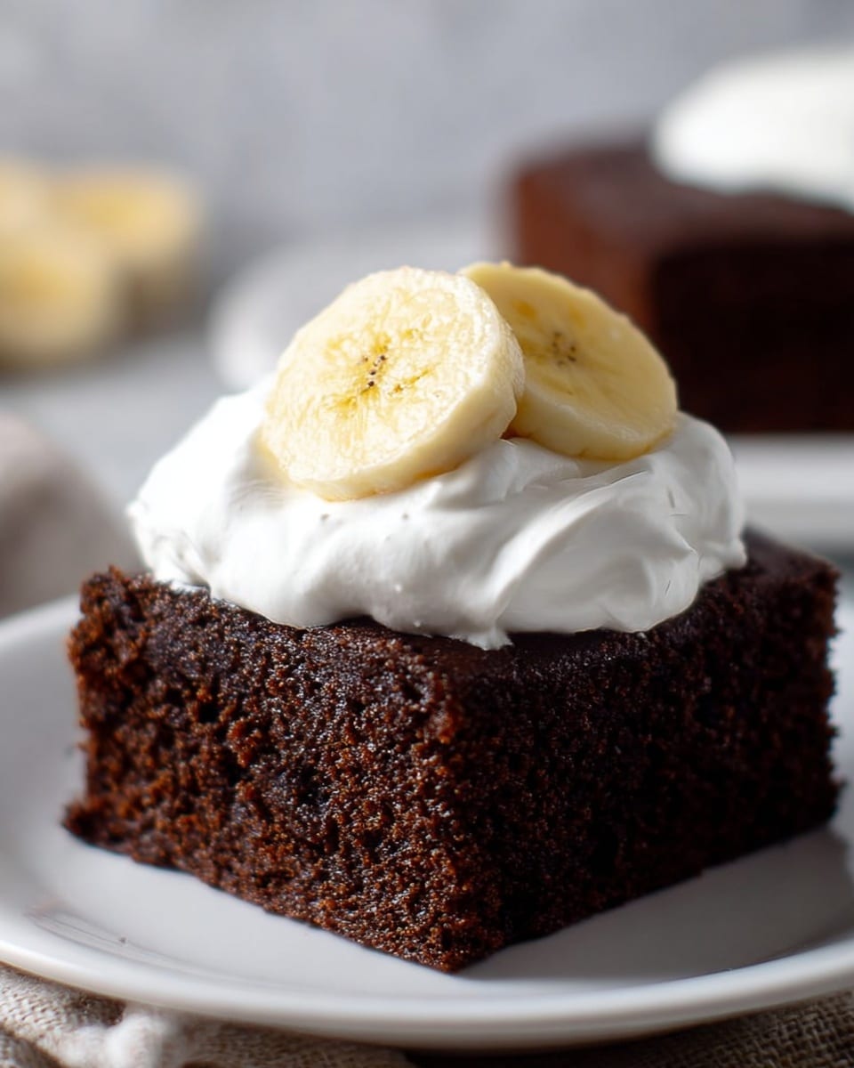A single square piece of dark brown chocolate cake with a moist and slightly crumbly texture sits centered on a white plate. On top of the cake is a thick dollop of smooth white whipped cream, which slightly spills over the edges of the cake. Resting on the whipped cream are two thick slices of pale yellow banana, positioned side by side. The background shows a soft white marbled texture and a blurred second piece of cake, creating a simple and clean setting. photo taken with an iphone --ar 4:5 --v 7