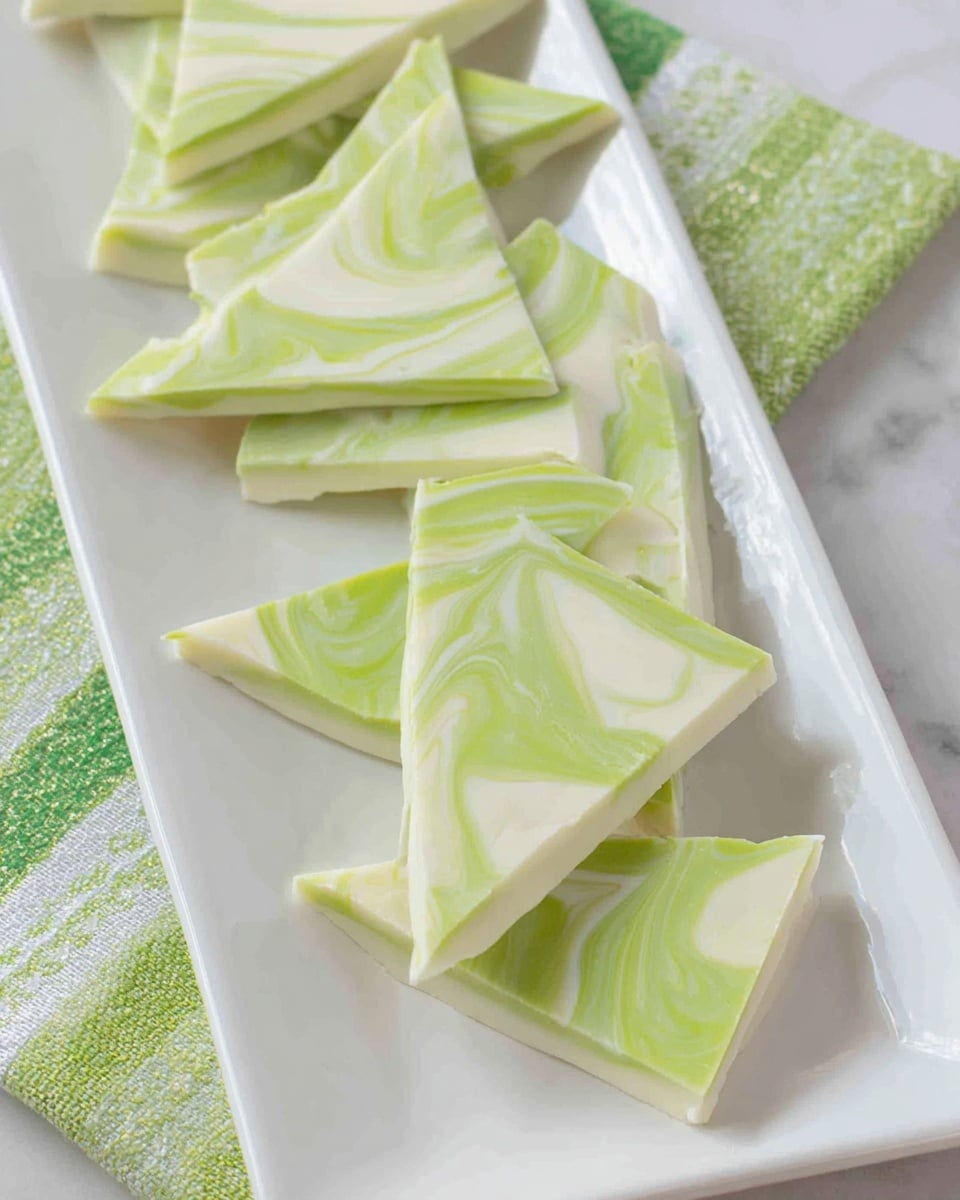 The image shows several pieces of marbled bark candy arranged on a long white rectangular plate. Each piece has two layers: the bottom layer is smooth and white, while the top layer features a light green and white swirl pattern, giving a soft, creamy texture. The pieces are irregularly shaped, with sharp edges and flat surfaces. The plate rests on a white marbled surface with a green and white striped cloth nearby. Photo taken with an iphone --ar 4:5 --v 7