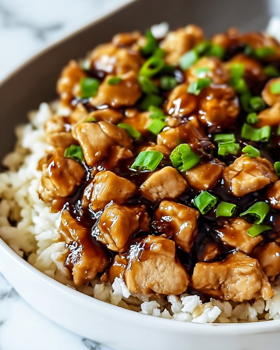 The image shows a close-up of a dish on a white plate with a curved edge. The bottom layer consists of white cooked rice with visible grains. On top of the rice, there is a generous layer of bite-sized chicken pieces covered in a thick, shiny brown sauce. Scattered over the chicken are small chopped green onions adding bright green color. The sauce looks glossy and sticky, giving a fresh and rich texture to the dish. The background is a white marbled texture. photo taken with an iphone --ar 4:5 --v 7