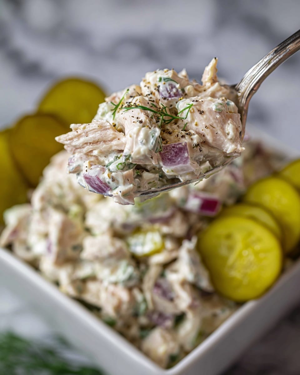 A close-up of a silver spoon holding a creamy chicken salad with visible chunks of white chicken meat mixed with small pieces of purple onion, green herbs, and black pepper flakes. The spoon is held above a white square bowl filled with the same chicken salad, and there are several light yellow-green pickle slices placed at the back edge of the bowl. The background shows a white marbled texture. photo taken with an iphone --ar 4:5 --v 7