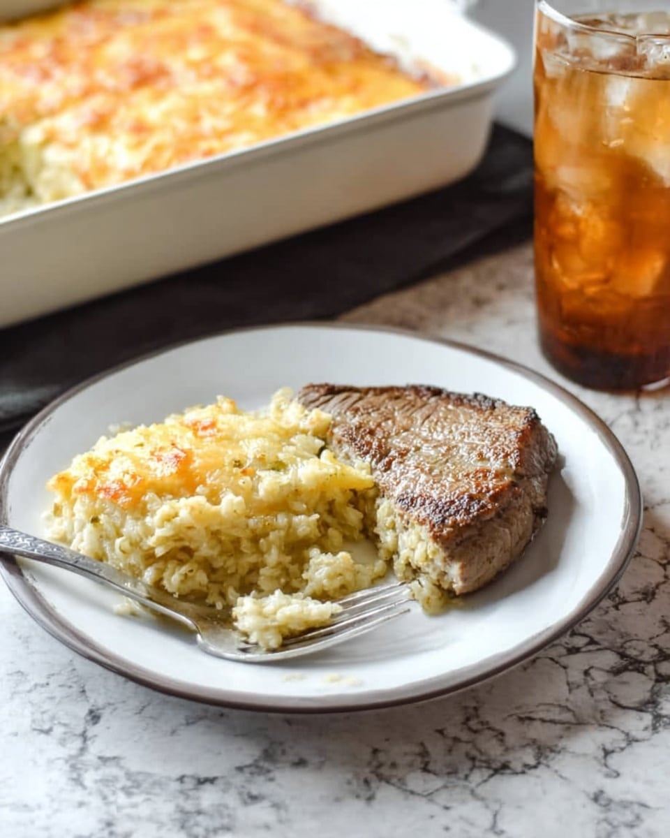 A white plate contains two main items: on the right side, there is a piece of cooked steak with a brown crust and slightly textured surface, and on the left side, a scoop of a baked, layered dish with a pale yellow creamy top and a grainy, light golden bottom layer. A silver fork rests on the left side of the plate, partially inserted into the baked dish. In the background, there is a rectangular white baking dish with more of the layered dish inside, showing a golden brown top. To the right, a clear glass filled with iced drink sits on a white marbled surface. Photo taken with an iphone --ar 4:5 --v 7