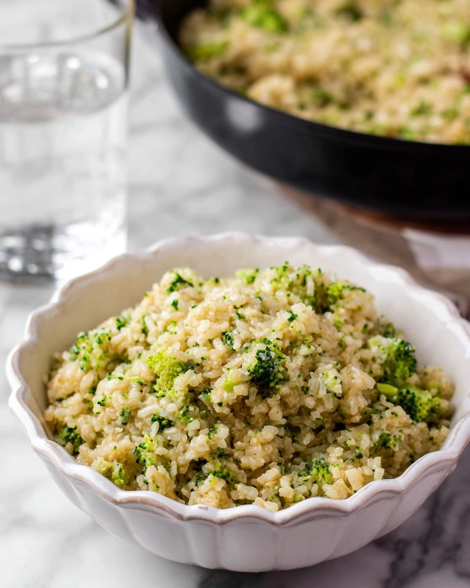 A close-up view of a creamy broccoli rice mixture piled high in a white bowl with decorative edges, showing light beige grains of rice mixed with small bright green broccoli bits creating a textured, soft appearance; behind the bowl is a black pan filled with more of the same broccoli rice and a clear glass of water, all placed on a white marbled surface. photo taken with an iphone --ar 4:5 --v 7