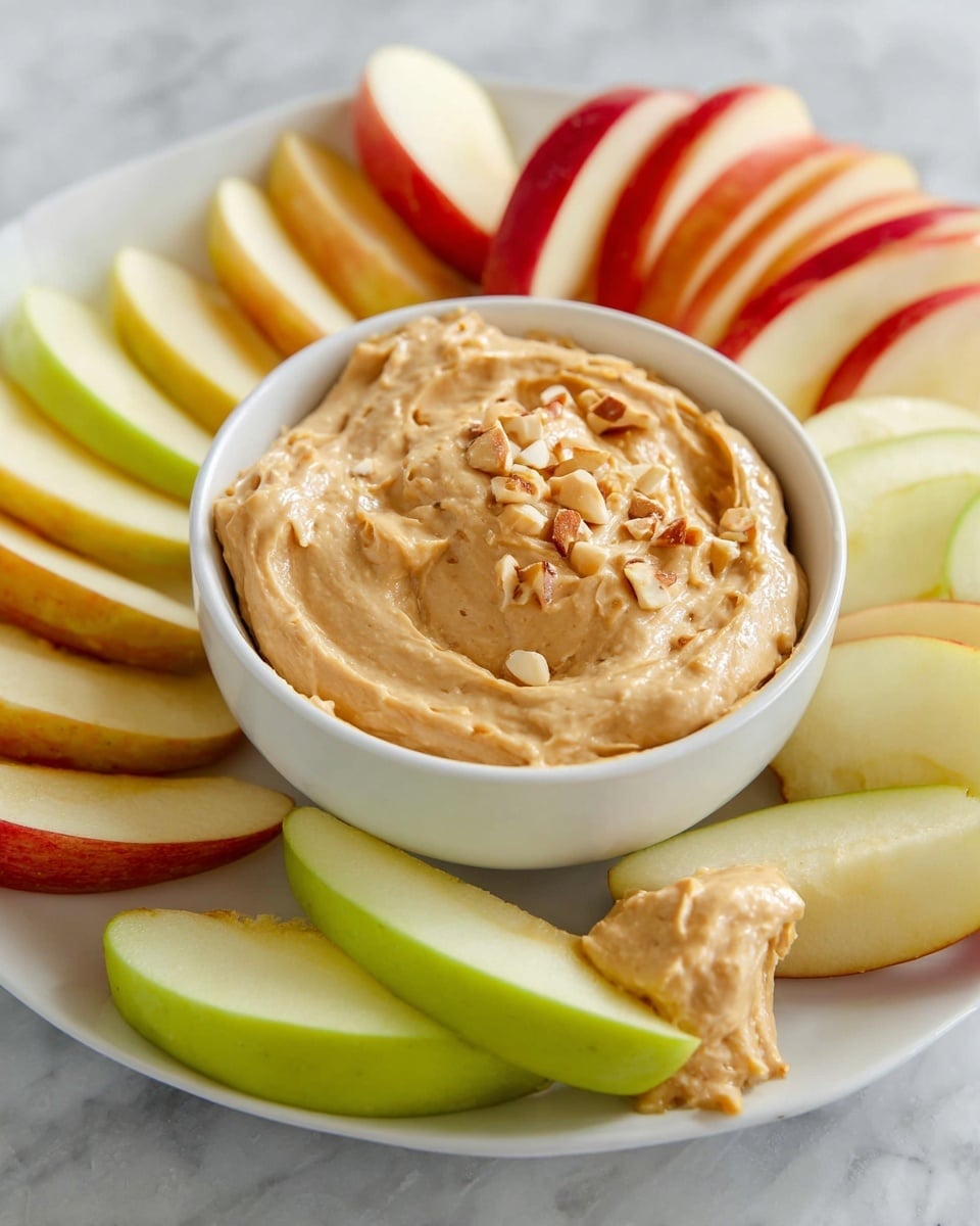 A white plate with curved edges holds three types of apple slices arranged in separate sections: green apple slices with a smooth, bright green peel and white flesh; red apple slices with a shiny, red and yellow peel and creamy white flesh; and yellow apple slices with a yellow-orange peel and pale flesh. In the middle of the plate, a small white bowl contains creamy peanut butter dip with a thick, slightly chunky texture, topped with small peanut pieces. Small green herb sprigs are placed near the apple slices on the plate, all set on a white marbled surface. Photo taken with an iphone --ar 4:5 --v 7