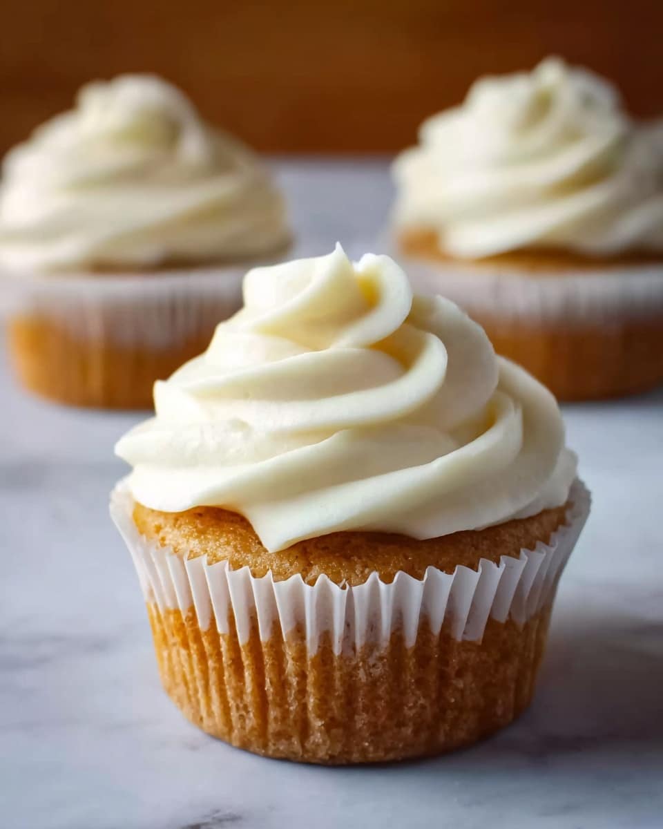 The image shows a close-up of a cupcake with three visible layers: the bottom layer is a golden-brown cake with a soft texture held in a white paper wrapper with ruffled edges; on top is a thick layer of smooth, creamy off-white frosting, swirled in a spiral shape that ends in a small peak at the center. In the background, two more cupcakes with the same layers and frosting are slightly blurred, placed on a white marbled surface. photo taken with an iphone --ar 4:5 --v 7