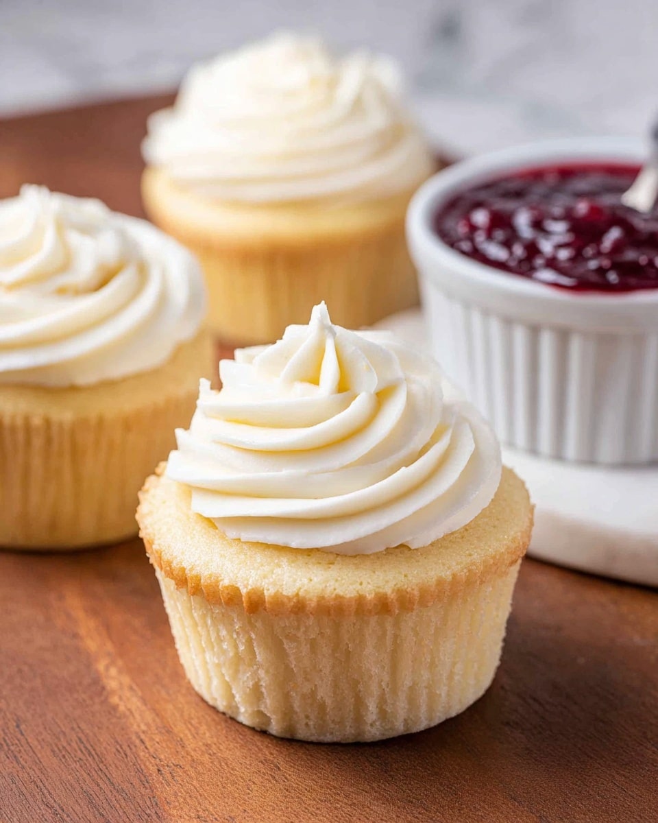 A light yellow vanilla cupcake with a smooth texture is topped with a swirl of thick, creamy white frosting that forms a small peak in the center. Behind it, two more vanilla cupcakes with the same thick white frosting are slightly out of focus. To the right, a white ramekin filled with a shiny dark red fruit jam sits on a white marbled surface. The cupcakes are resting on a wooden surface that contrasts softly with their light color. photo taken with an iphone --ar 4:5 --v 7
