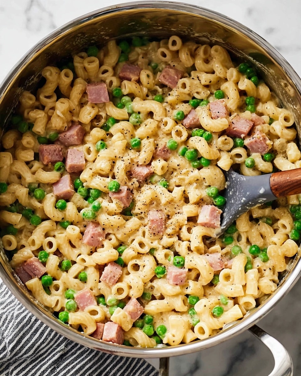 A close-up view of a shiny silver pot filled with creamy pasta made of small, ring-shaped noodles, mixed evenly with bright green peas and small pinkish-brown cubes of cooked ham, all coated in a light beige sauce and sprinkled with ground black pepper. A dark gray spoon with a wooden handle is placed inside the pot on the right side, resting against the pasta. The pot sits on a white marbled surface with a striped cloth partially visible under the handle. photo taken with an iphone --ar 4:5 --v 7