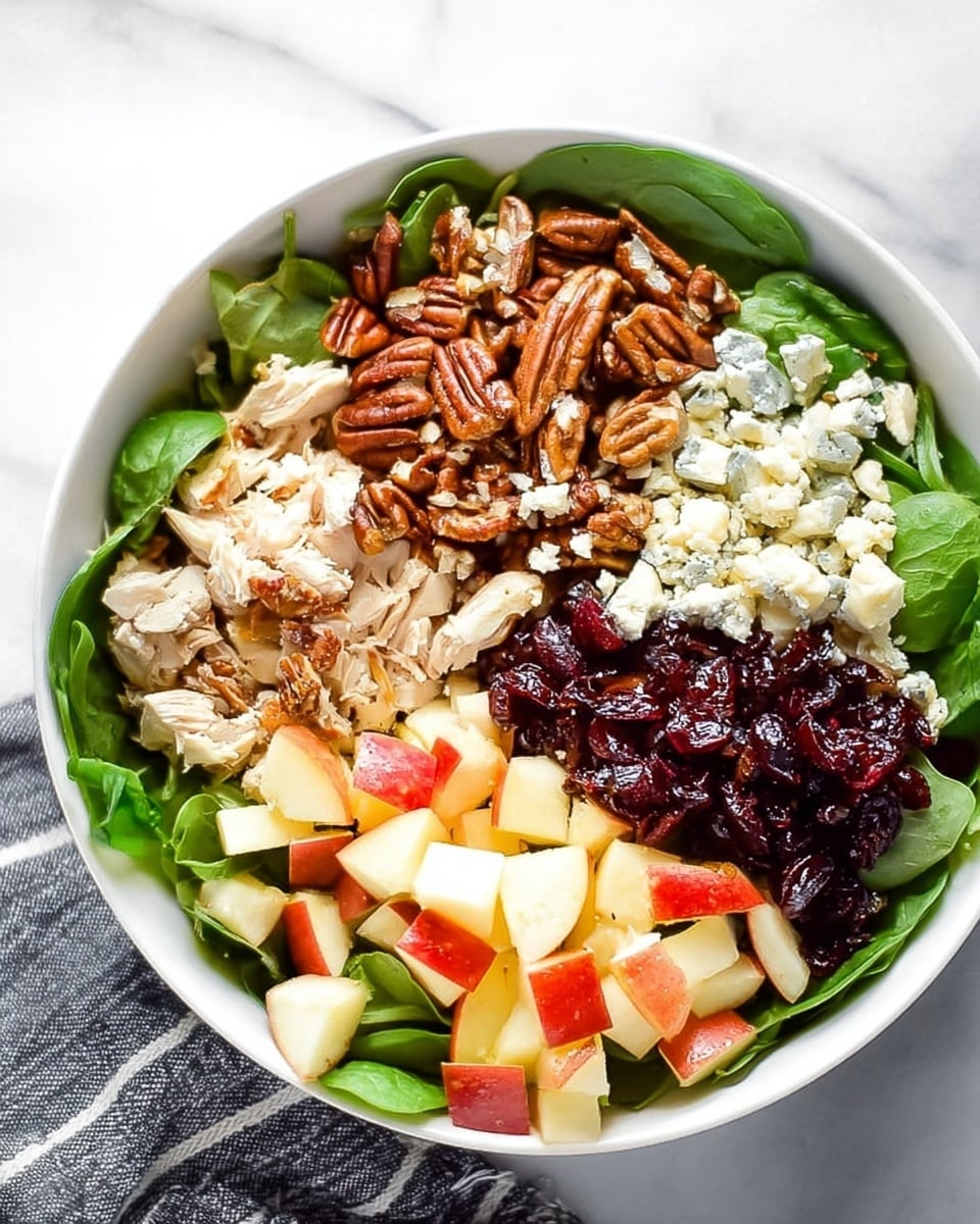 A white bowl filled with a fresh spinach salad divided into sections on top: bright green spinach leaves form the base layer, topped with chopped roasted pecans that are shiny and brown, small white crumbles of soft cheese scattered over pieces of shredded pale chicken, diced red and yellow apple cubes showing their skin and flesh, and a mound of dark red dried cranberries in the center. The colors contrast well with the white bowl and the salad looks fresh and textured. It is placed on a white marbled surface with a striped gray and white cloth nearby. Photo taken with an iphone --ar 4:5 --v 7