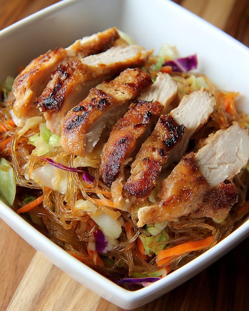 The image shows a white rectangular bowl filled with a layer of thin, cooked glass noodles mixed with cooked shredded vegetables like orange carrot strips and light green and purple cabbage pieces, creating a colorful and slightly shiny base. On top, there are six pieces of sliced grilled chicken breast, arranged in a row, showing a golden brown, slightly crispy skin with charred marks and juicy white meat inside. The bowl is placed on a wooden surface. photo taken with an iphone --ar 4:5 --v 7
