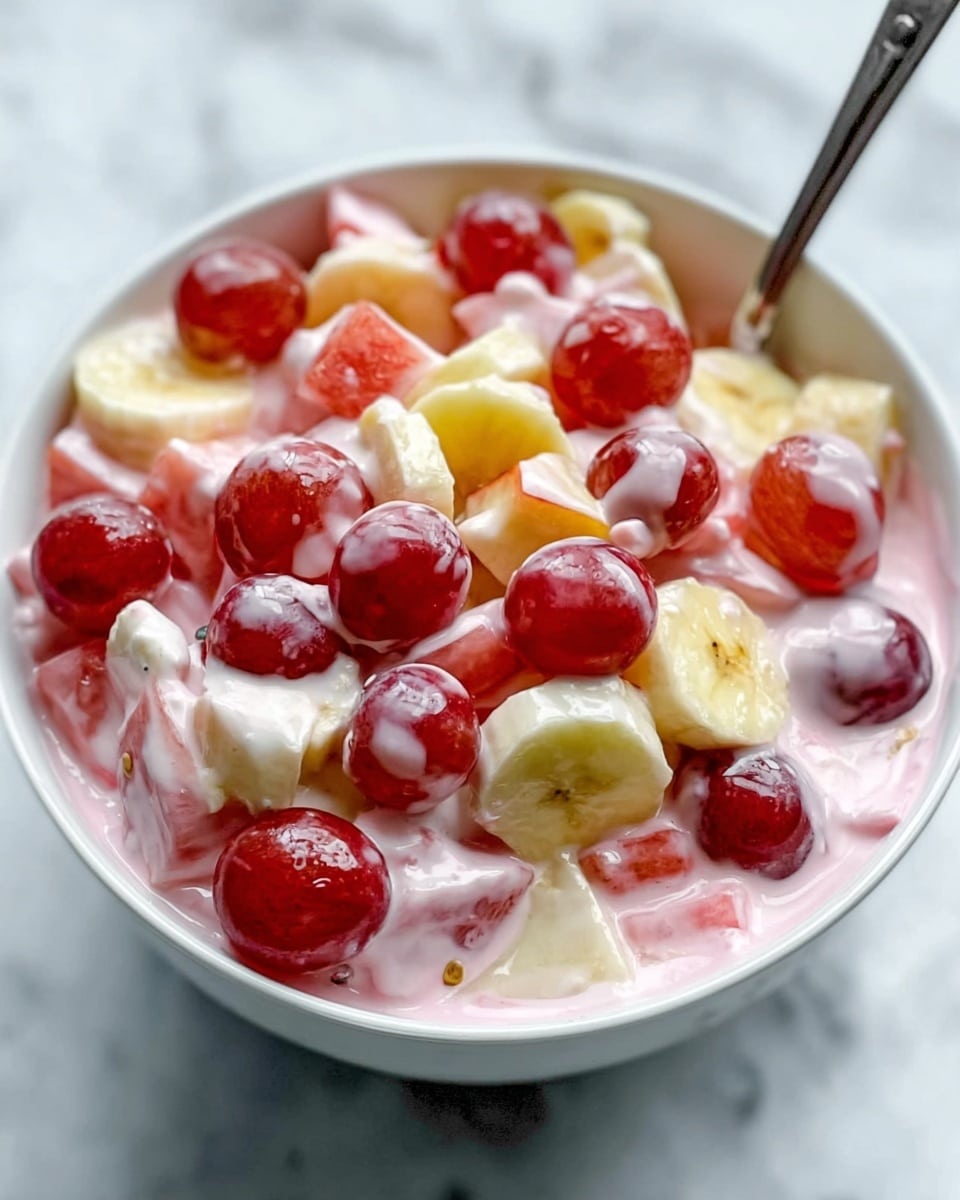 A close-up view of a white bowl filled with a creamy fruit salad that has three main layers. The bottom layer is a smooth, pink yogurt or cream that covers the base. The middle layer is a mix of small fruit pieces, including light yellow banana slices, pale beige apple chunks, and bright red grapes. The top layer consists of round red grapes scattered over the creamy mixture. A shiny silver spoon is inserted into the bowl. The bowl sits on a white marbled surface. Photo taken with an iphone --ar 4:5 --v 7