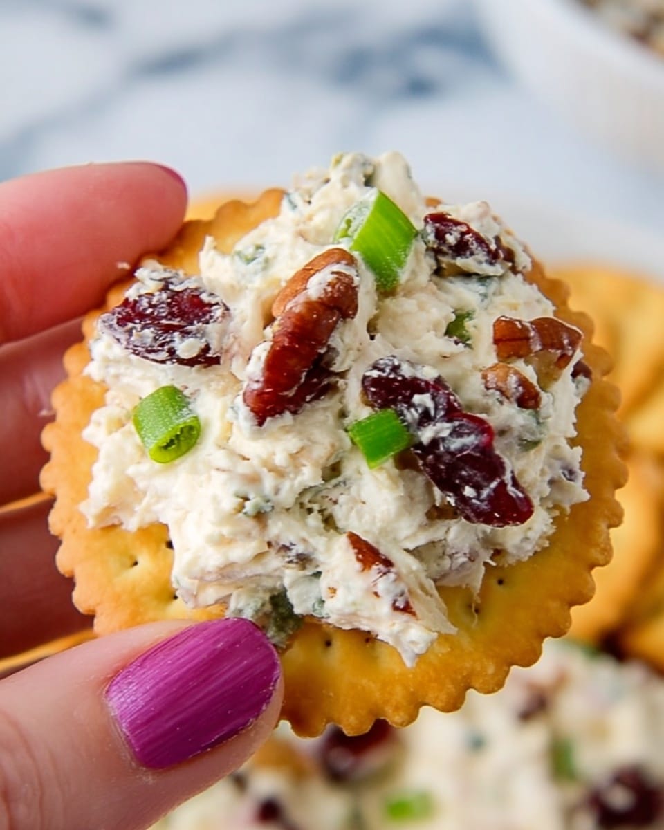 A close-up image shows a woman's hand holding a round, light golden cracker with scalloped edges. On the cracker, there is a thick scoop of creamy white cheese mixed with small chunks of pecans, bright green pieces of chopped green onion, and dark red dried cranberries. The creamy cheese mixture looks soft and textured, contrasting with the crunchy cracker. The background features a white marbled surface, softly blurred to keep the focus on the cracker and topping. photo taken with an iphone --ar 4:5 --v 7