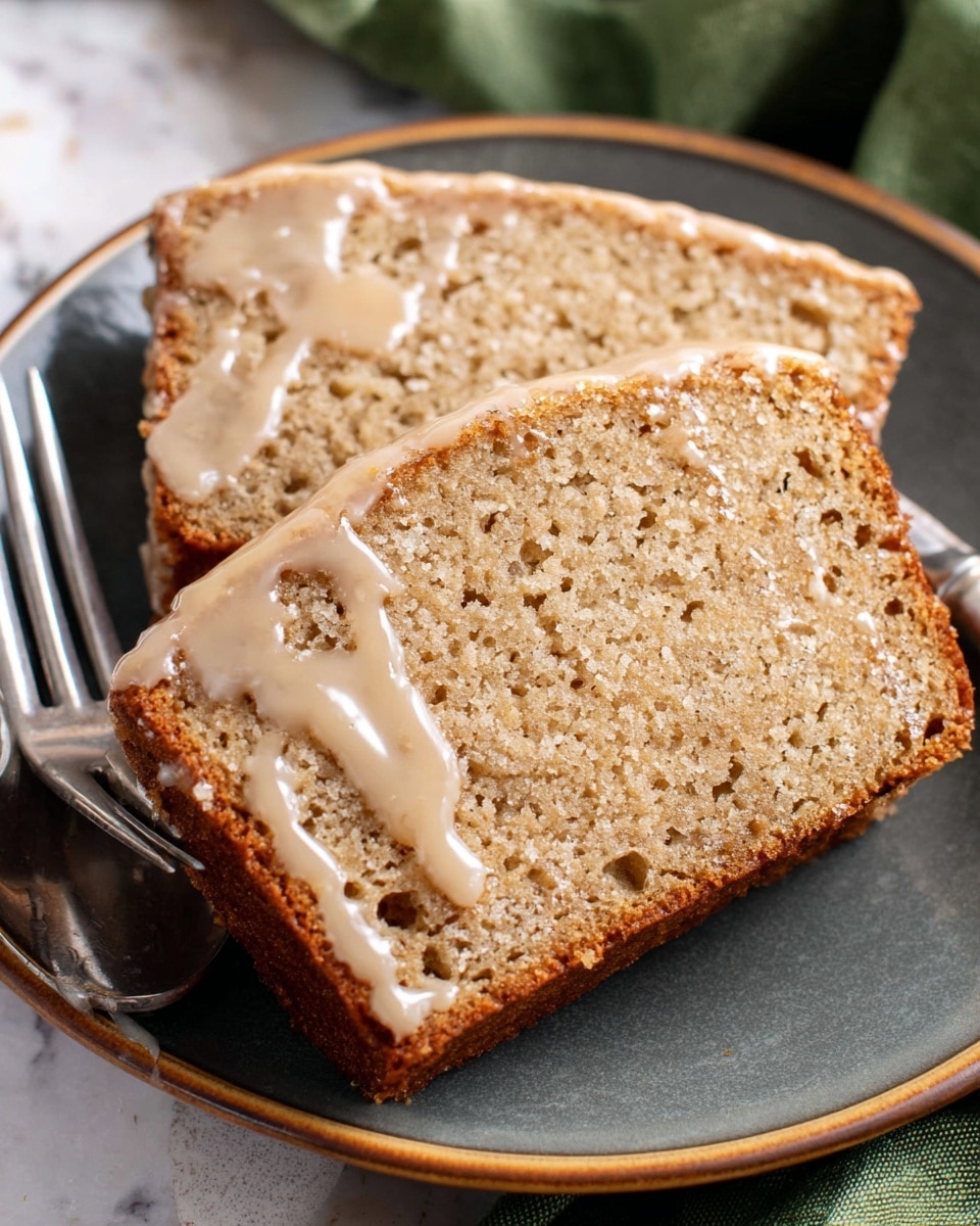 Two slices of light brown moist bread loaf with a soft and spongy texture sit stacked slightly on a white plate with a dark gray finish. The bread has visible tiny air holes and a shiny glaze of light beige icing drizzled unevenly over the top, dripping slightly down the sides. A silver fork and spoon rest beside the slices on the plate. The plate is set on a surface with a white marbled texture, and a green fabric is folded loosely in the background. Photo taken with an iphone --ar 4:5 --v 7
