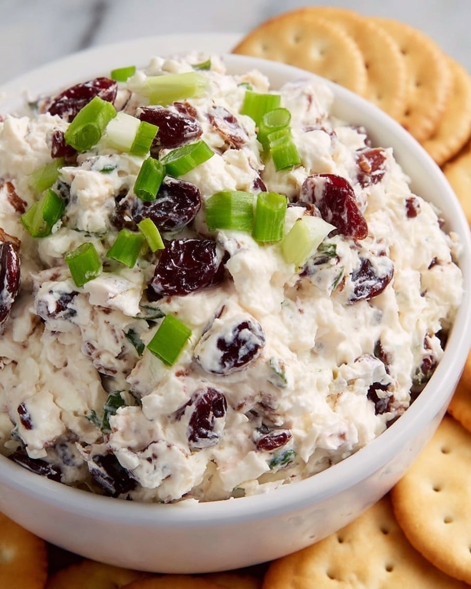 A close-up view of a bowl filled with a creamy white cheese mix layered with small dark raisins and deep red dried cranberries, topped with bright green slices of fresh green onion. The texture looks soft and slightly chunky, showing the mix of ingredients evenly spread throughout. The bowl is white, sitting on a white marbled surface, with light golden round crackers arranged neatly beside it. Photo taken with an iphone --ar 4:5 --v 7