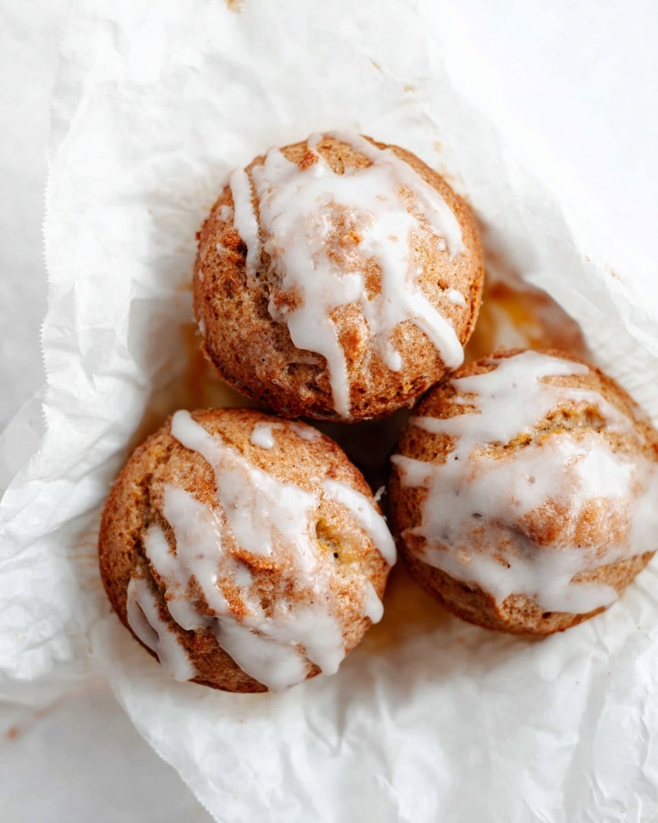A close-up top view of three round muffins baked together in white parchment paper. The muffins have a golden-brown color with a slightly rough texture on top. They are drizzled with a shiny white glaze, mostly covering the center of each muffin, with some glaze flowing between them. The background is a clean white marbled texture giving a soft and bright look to the image. Photo taken with an iphone --ar 4:5 --v 7