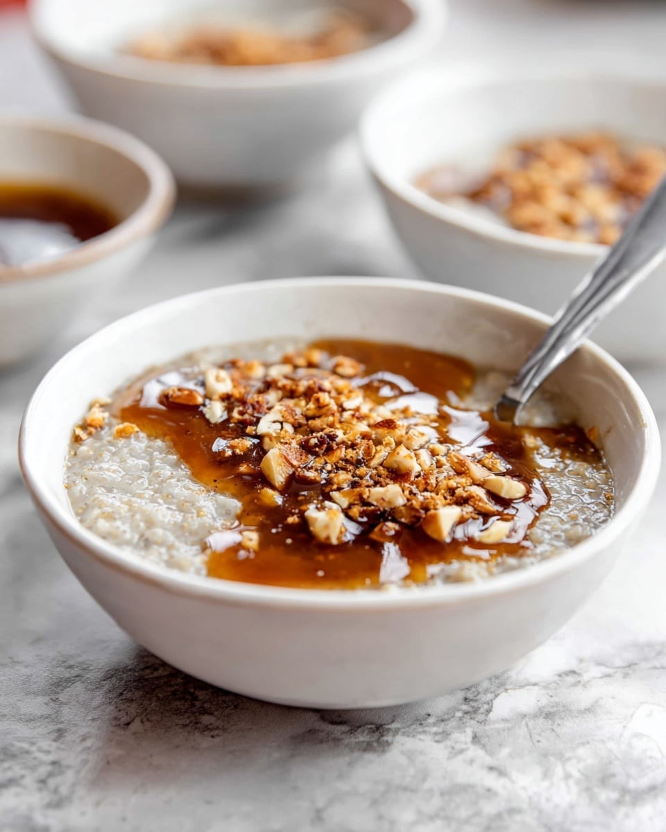 A white bowl filled with a thick, light gray porridge in the base layer, topped with a shiny dark brown syrup layer that pools unevenly on the surface, sprinkled with crushed brown nuts. A silver spoon rests inside the bowl on the right side. In the soft-focus background, two more white bowls hold the same porridge mixture and nut topping. The setting has a white marbled surface underneath, giving a clean and bright look. Photo taken with an iphone --ar 4:5 --v 7