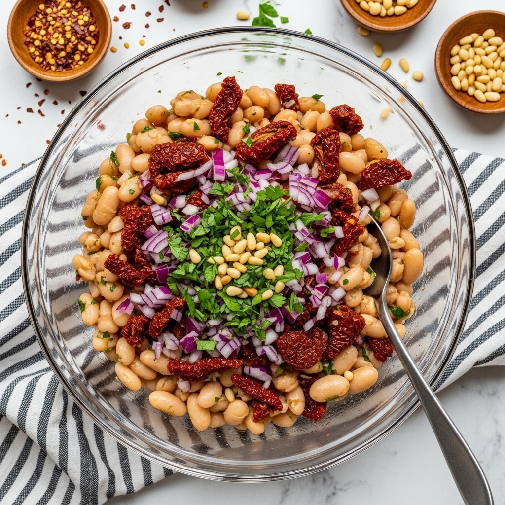 A clear glass bowl filled with a vibrant three-layer mix: the base is creamy white cannellini beans, scattered throughout with chunky pieces of dark red sun-dried tomatoes and finely chopped purple-red onions, all topped with fresh chopped green herbs and small golden pine nuts. A spoon is placed inside the bowl, ready to serve. The bowl sits on a white marble surface with a black and white striped cloth partially beneath it. Around the bowl, small wooden dishes hold red pepper flakes and more pine nuts. Photo taken with an iphone --ar 4:5 --v 7