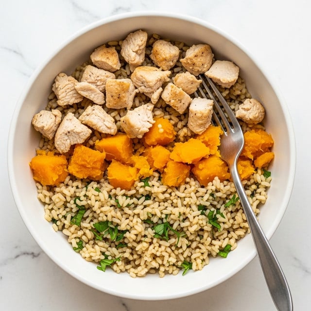A white bowl is filled with a rice dish that has three main layers visible: the bottom layer is light brown rice mixed with small pieces of green herbs; the middle layer includes soft chunks of orange pumpkin or squash; the top layer has pieces of light brown cooked chicken scattered evenly. A silver fork rests inside the bowl, angled slightly to the right. The background is a white marbled surface. The photo taken with an iphone --ar 4:5 --v 7