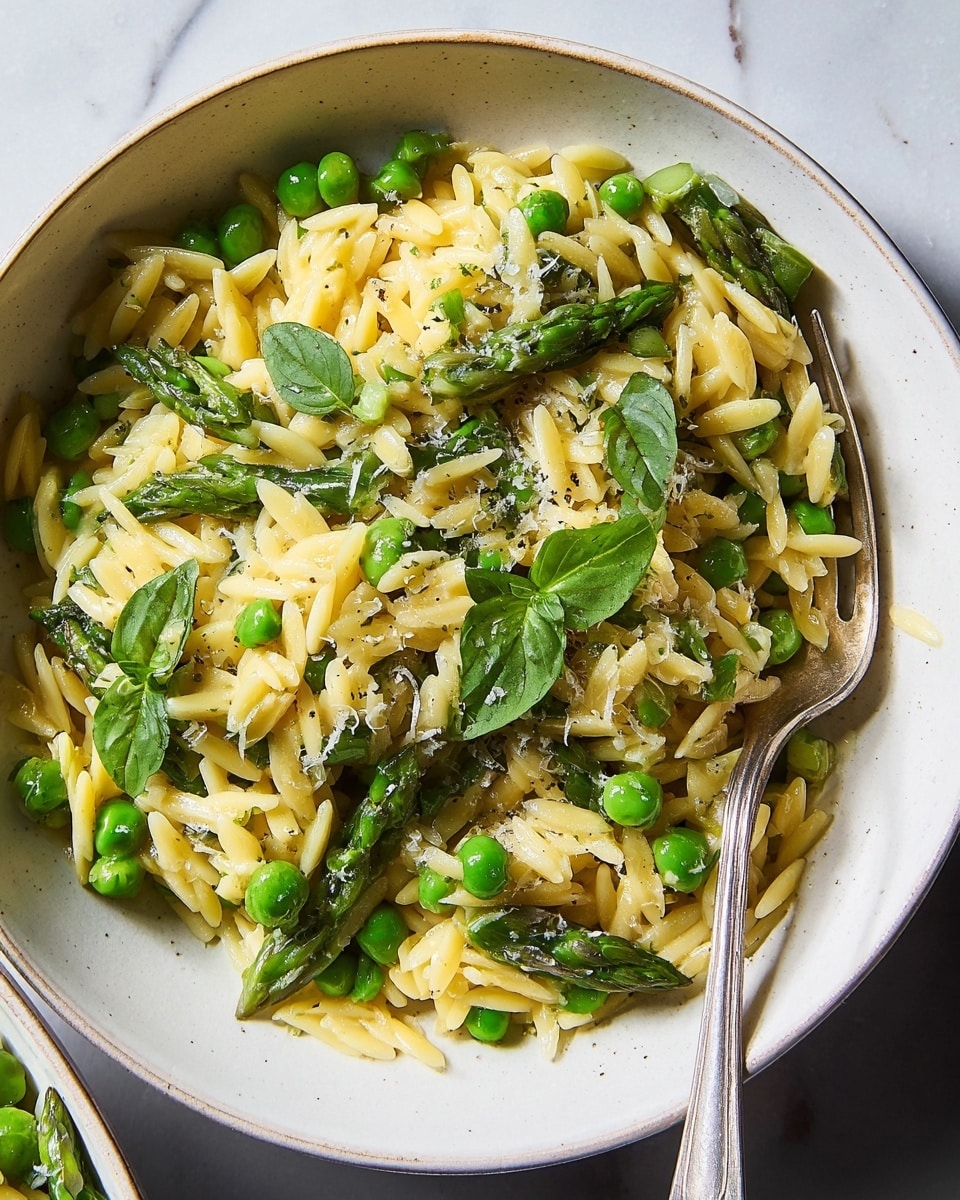 A close-up of a white bowl filled with a single layer of cooked orzo pasta mixed with bright green peas and cut asparagus pieces scattered evenly, topped with fresh green leaves and lightly grated cheese, all lightly coated in a glossy sauce with visible black pepper specks. A silver fork is placed on the right side inside the bowl, resting against the edge, all set on a white marbled surface. photo taken with an iphone --ar 4:5 --v 7