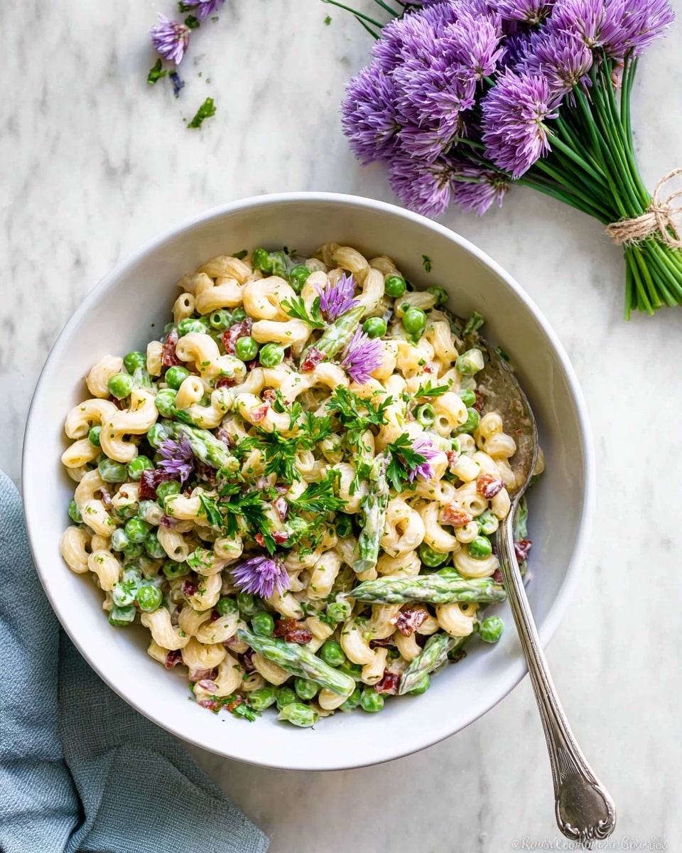 A white bowl filled with a creamy pasta salad made of three layers: small elbow macaroni in pale yellow, bright green peas and chopped asparagus pieces scattered evenly, and small bits of red sun-dried tomatoes mixed throughout. The salad is garnished with fresh green parsley and tiny purple edible flowers on top. A silver fork rests inside the bowl on the right side. The bowl and fork sit on a white marbled surface, with a small bunch of purple chive flowers tied with string placed in the upper right corner, and a light blue cloth partially visible in the lower left corner. photo taken with an iphone --ar 4:5 --v 7