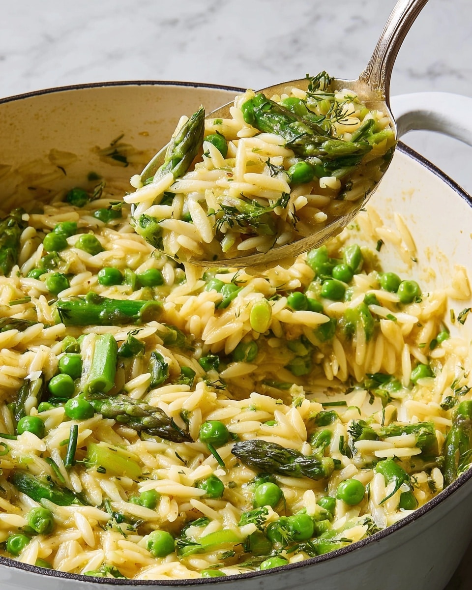 The image shows a close-up of a creamy yellow orzo pasta mixed with bright green peas and chopped asparagus pieces in a white pot. The orzo layer is soft and saucy, with the green vegetables peeking through evenly spread. A spoon lifts a portion of the dish from the pot, showing the texture of the pasta and the freshness of the green vegetables. The background is a white marbled texture. photo taken with an iphone --ar 4:5 --v 7